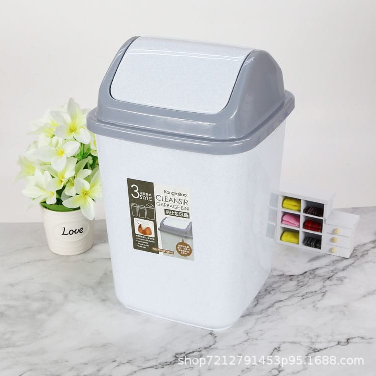 A white and gray plastic kitchen trash can with a hinged lid, shown on a marble surface next to a small potted flower and a small organizer tray. The front has a product label.