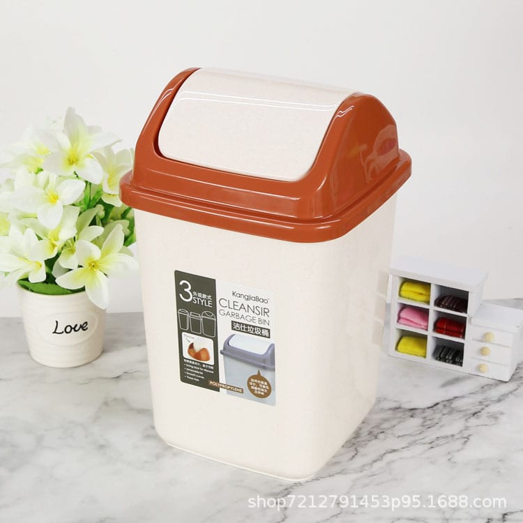 A plastic kitchen trash can with a cream-colored body and a terracotta orange flip-up lid, shown on a marble countertop with a flower pot and a small organizer in the background.