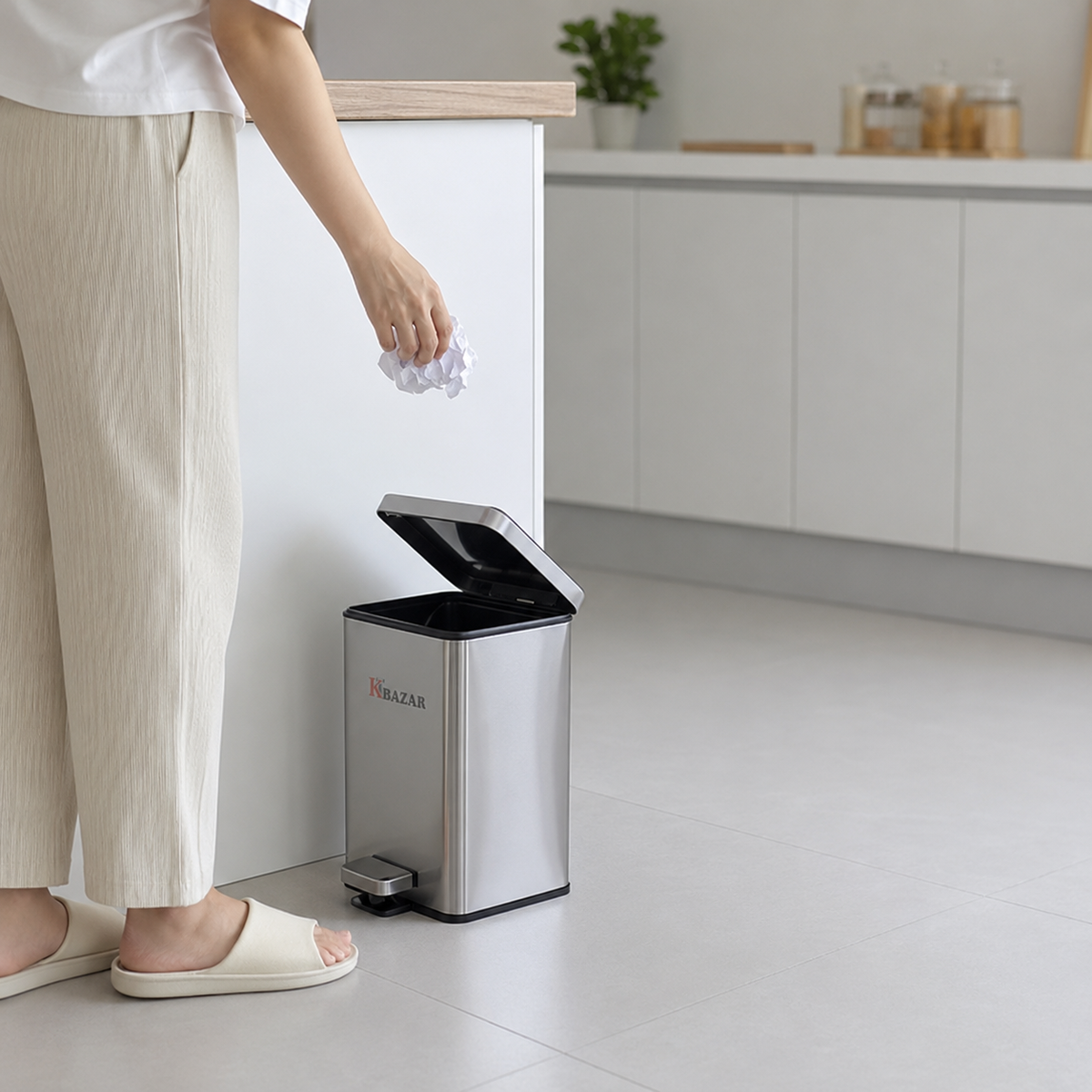 A person in a white top and beige pants is disposing of a crumpled paper into a stainless steel step-through trash can with a black lid in a modern kitchen setting.