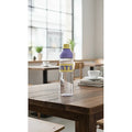 Clear bottle with a purple label on a wooden table in a room with large windows.