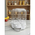A white and clear plastic multi-compartment food storage container with a transparent dome lid, placed on a marble countertop in front of wooden shelves filled with jars of pasta and grains.